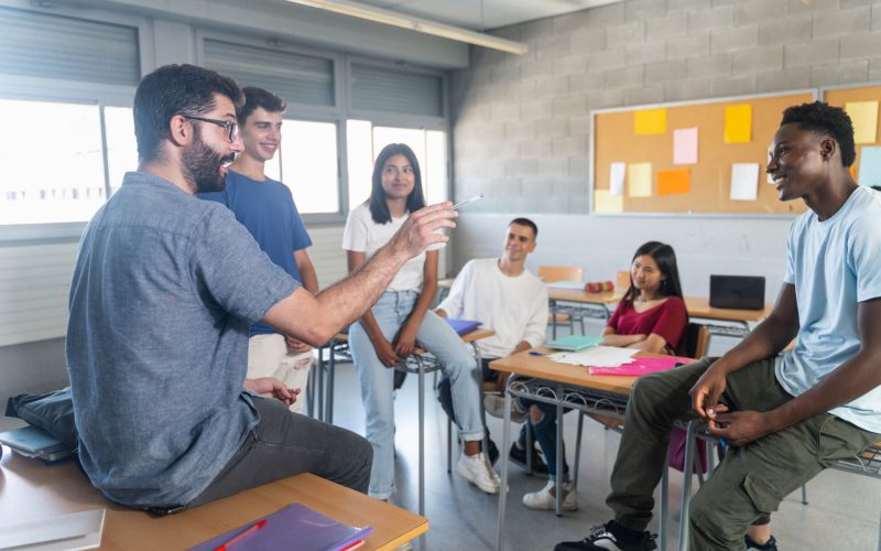 Group of teenager students with young teacher at classroom, sitting talking and sharing conversation together