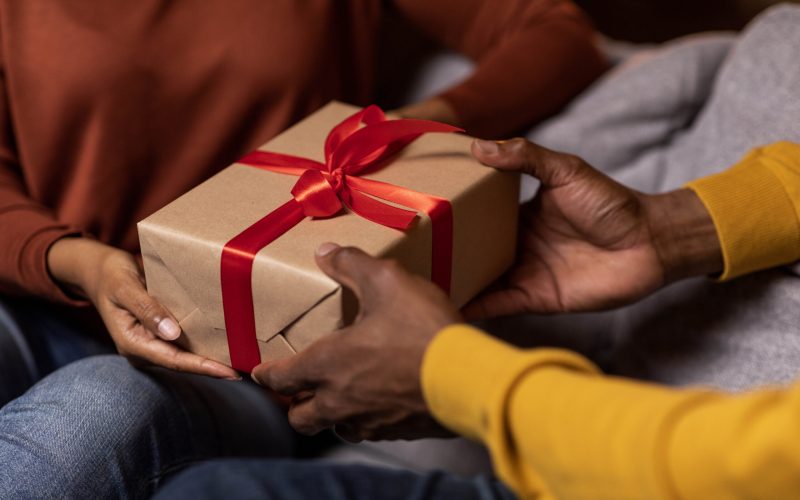 Cropped of black man and woman exchanging presents on anniversary day at home, unrecognizable couple holding gift box with red baw while sitting on couch together, having romantic evening