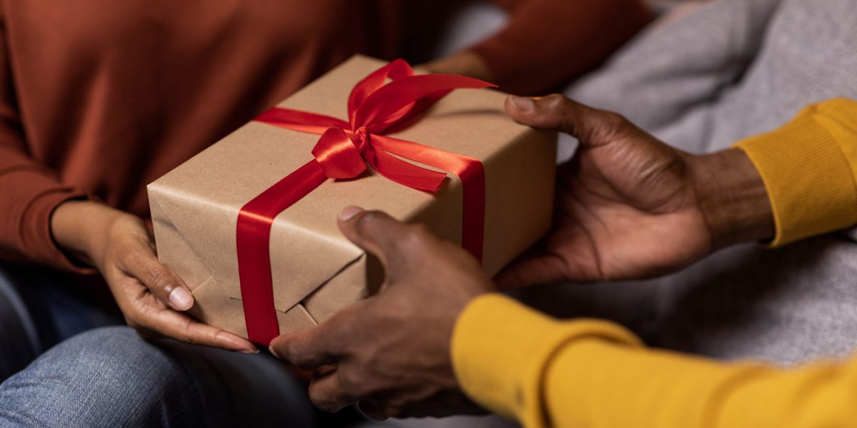 Cropped of black man and woman exchanging presents on anniversary day at home, unrecognizable couple holding gift box with red baw while sitting on couch together, having romantic evening
