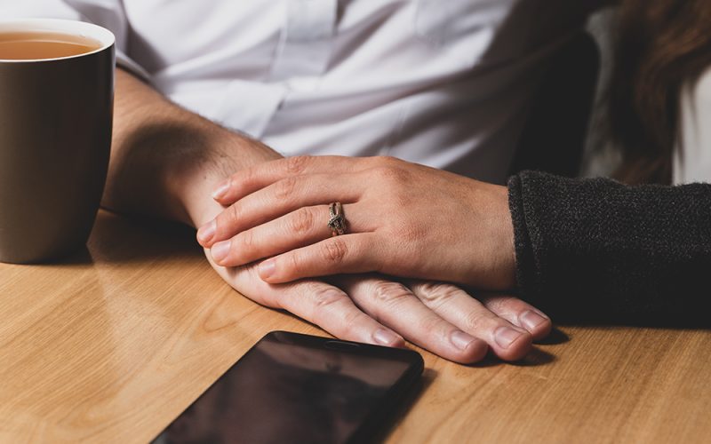 On a table the male hand lies, and from above it is caressed by a female hand. The female hand caresses a male hand. On a table phone lies and there is a cup of tea. The photo is tinted.
