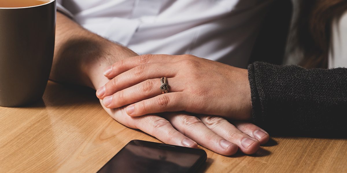On a table the male hand lies, and from above it is caressed by a female hand. The female hand caresses a male hand. On a table phone lies and there is a cup of tea. The photo is tinted.
