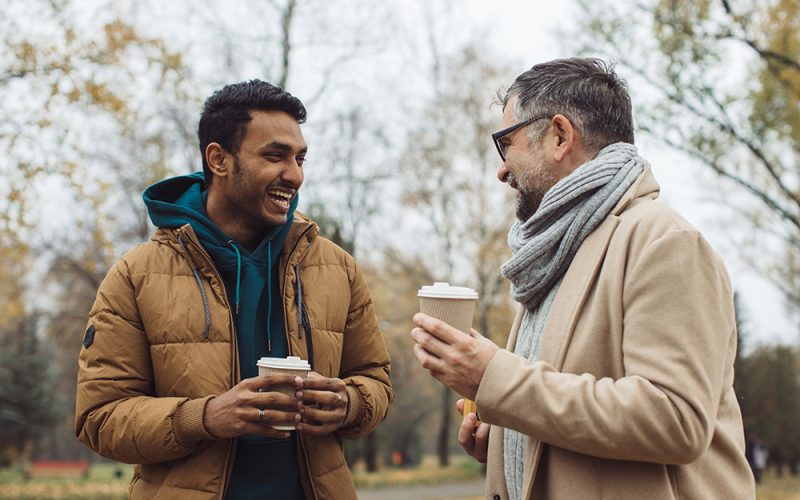 Friends, a senior and a young man walking and talking and drinking coffee together in the autumn park.