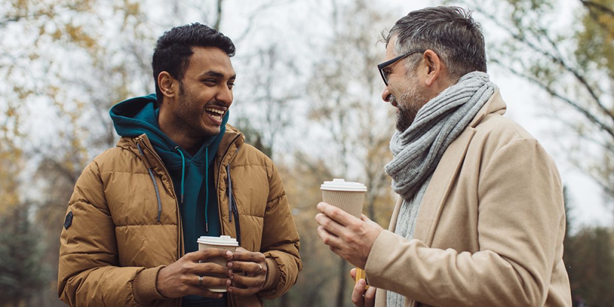 Friends, a senior and a young man walking and talking and drinking coffee together in the autumn park.
